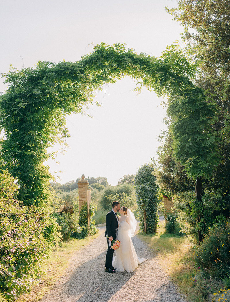 couple-portraits-villa-pozzolo-wedding-kissing-under-arch-13 Couple standing beneath ivy arch, while standing on a gravel road after their wedding at villa pozzolo in Montaione, Tuscany.