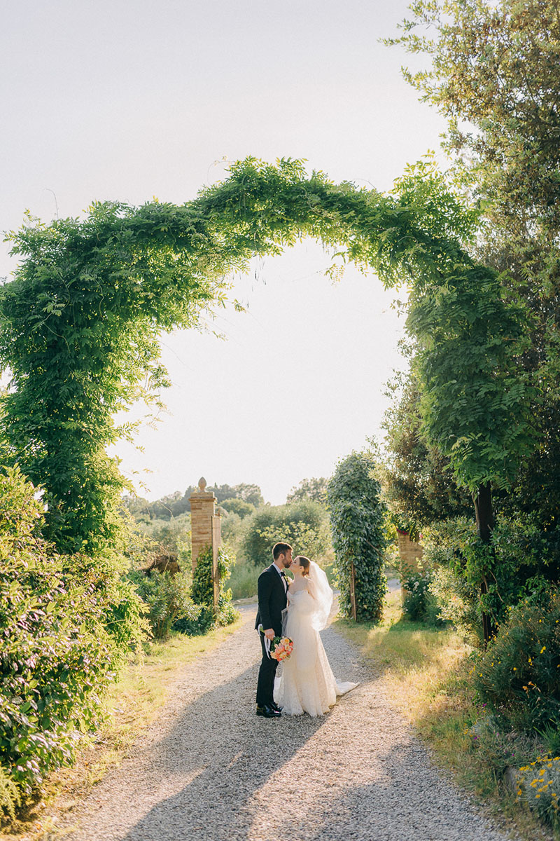 couple-portraits-villa-pozzolo-wedding-kissing-under-arch-13 Couple standing beneath ivy arch, while standing on a gravel road after their wedding at villa pozzolo in Montaione, Tuscany.