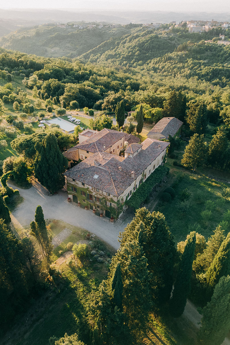hero-villa-pozzolo-wedding-aerial-view-02 Aerial photo of Villa Pozzolo surrounded by lush Tuscan hills, with the ivy-covered villa at center, terracotta roofs glowing in golden light, and cypress trees lining the gravel driveway.