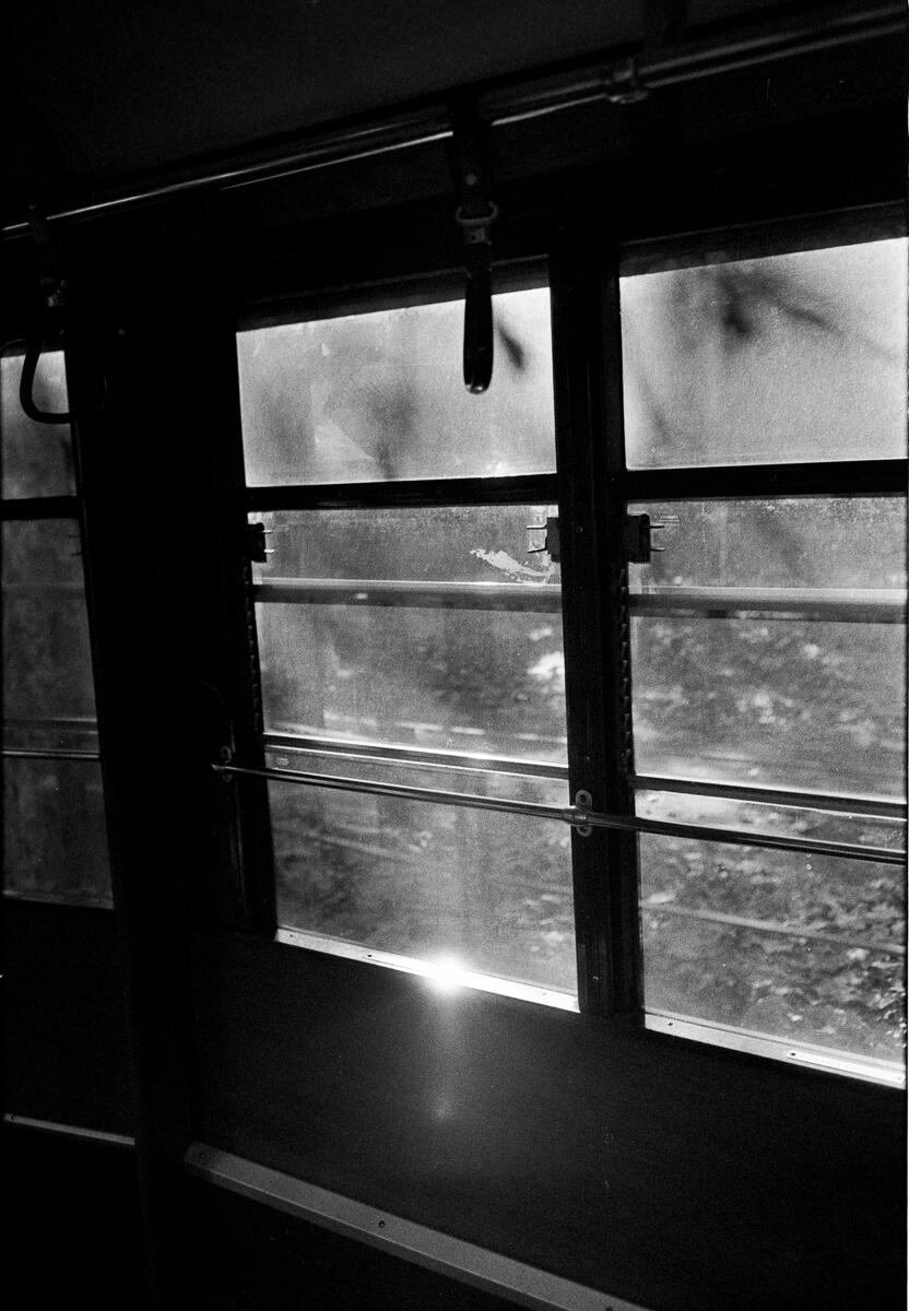 Black and white film photograph of a train carriage window, blurred landscape rushing past outside, strong contrast between the dark interior and light coming through the glass