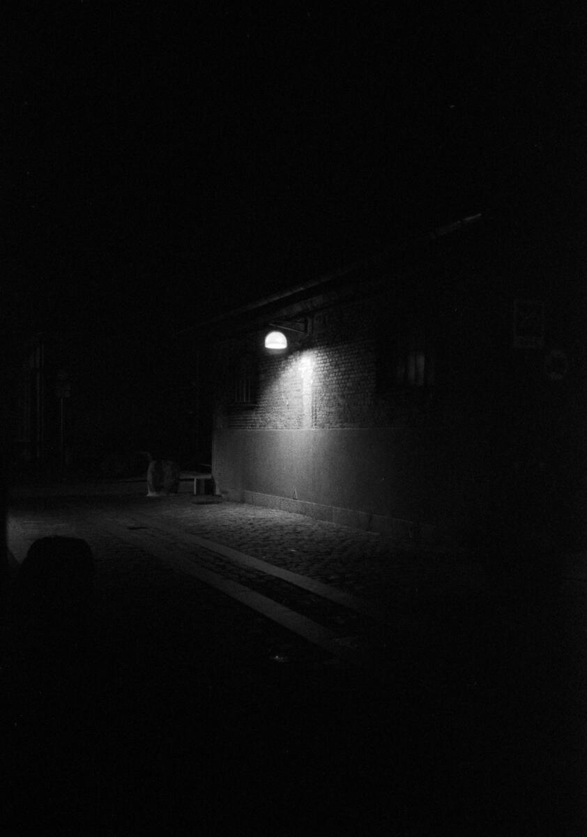 Black and white film photograph of a dark street at night, a single wall-mounted light illuminating a patch of brick building and cobblestoned pavement in deep darkness