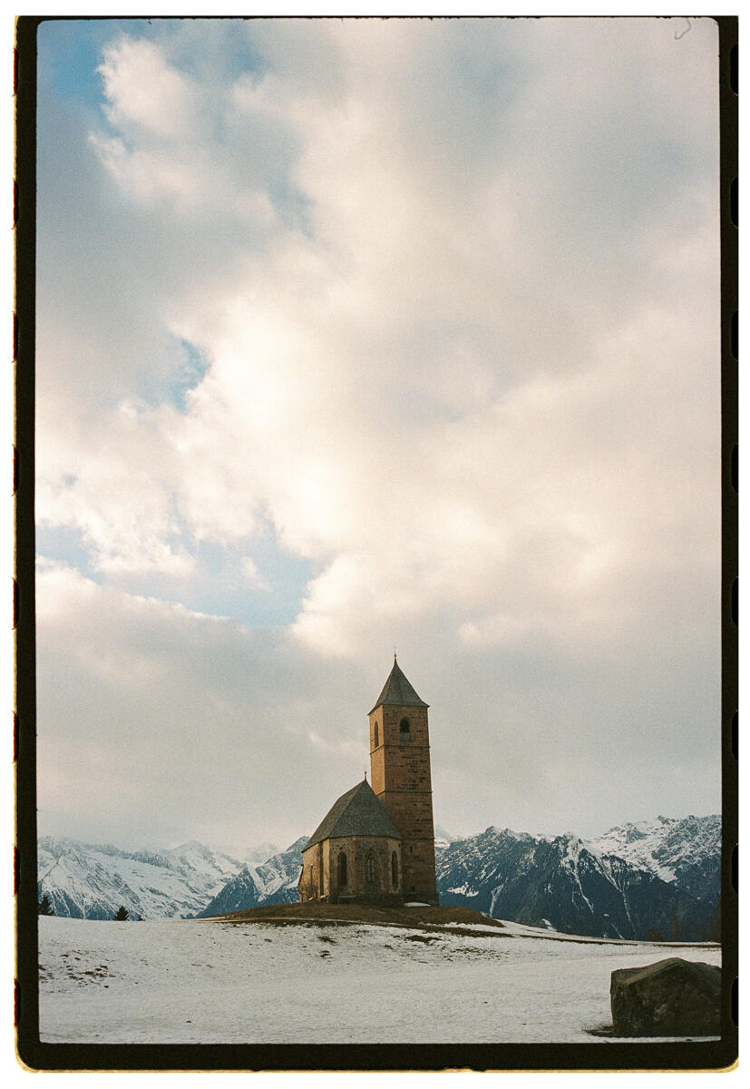 Film photograph of an isolated Romanesque stone church on a snow-covered hilltop, mountains in the background beneath a dramatic cloudy sky