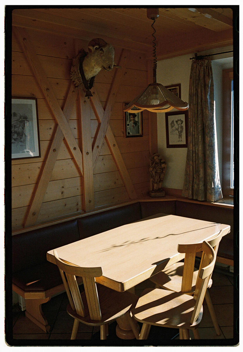 Film photograph of a mountain hut interior, sunlight falling across a wooden table and two chairs, hunting trophy mounted on the wall, framed prints and a figurine visible in the corner