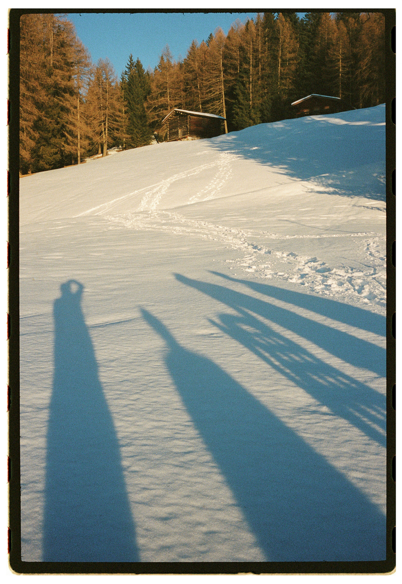 Film photograph of long afternoon shadows cast across a snowy alpine slope, the shadow of a person with a camera visible in the foreground, wooden chalets and larch trees in the background