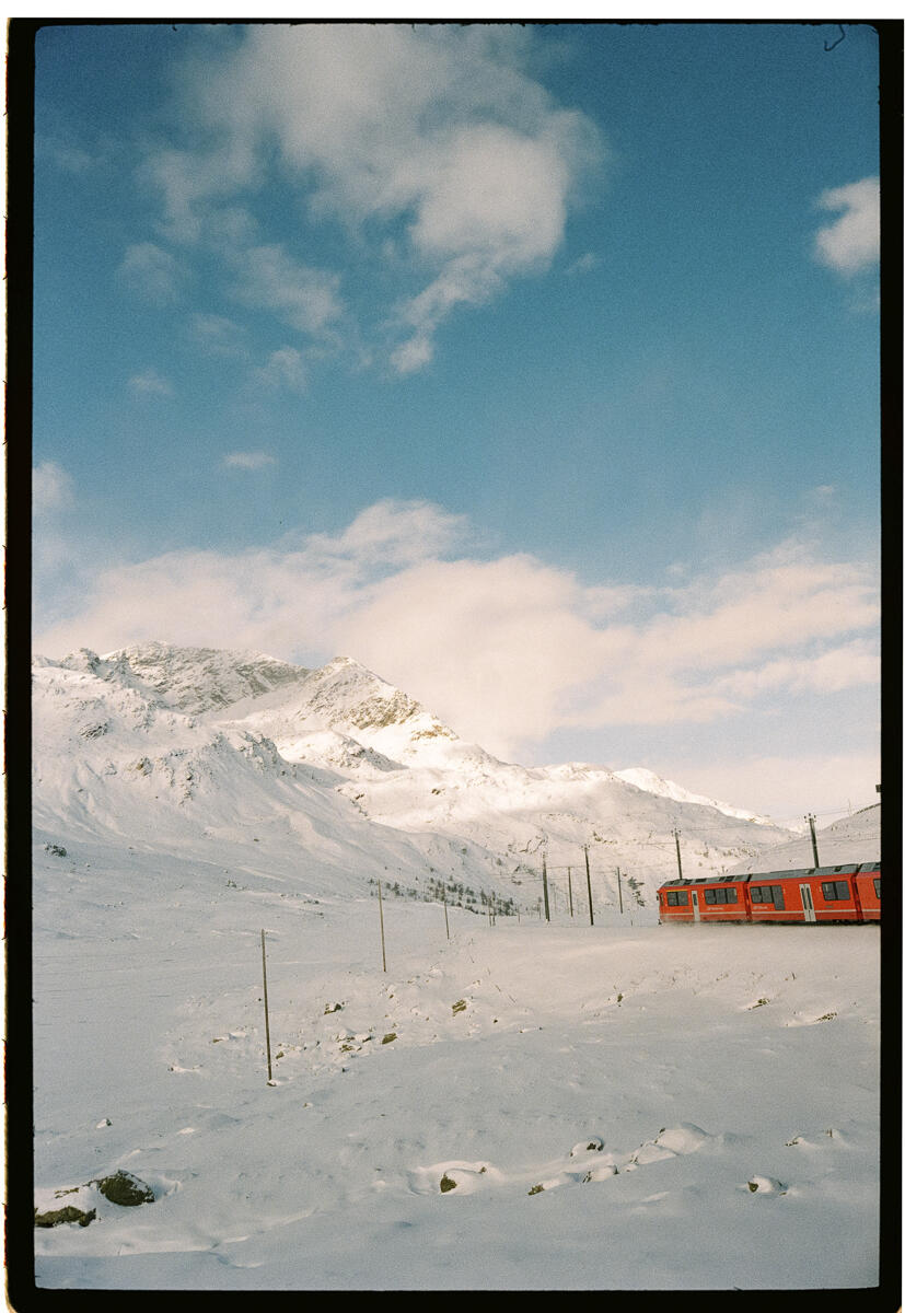 Film photograph of a red mountain train passing through a snow-covered alpine landscape, snowy peaks and blue sky with clouds in the background