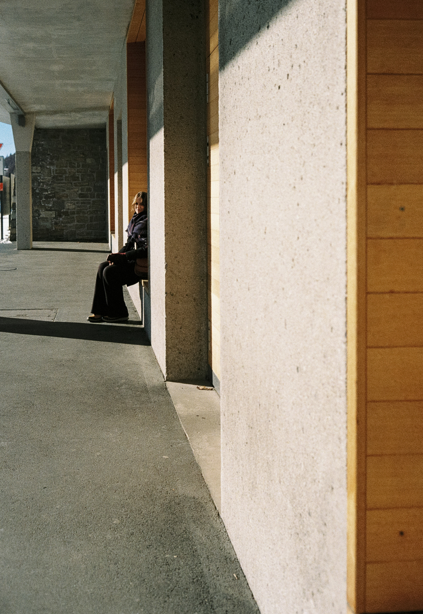 Film photograph of a woman sitting alone on a bench in a covered walkway, partially framed by a concrete column, sunlight falling across the pavement in an alpine town