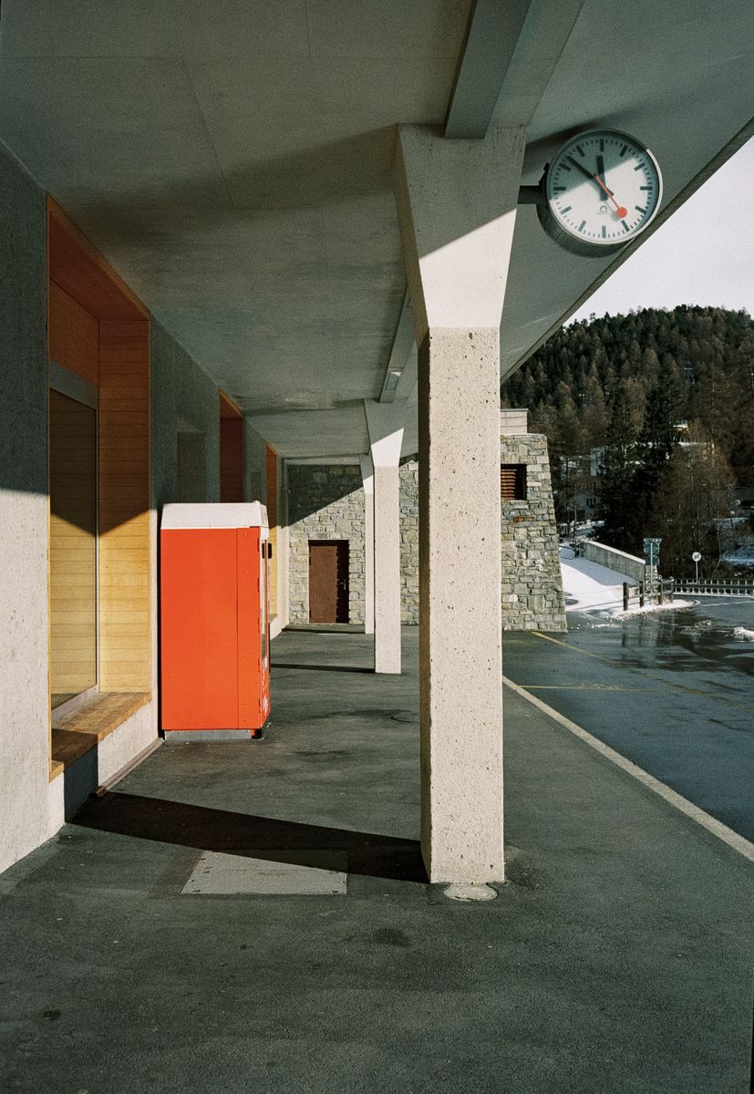 Film photograph of a covered railway station platform, an orange vending machine beside a concrete pillar, Swiss station clock on the right, mountains visible beyond the car park