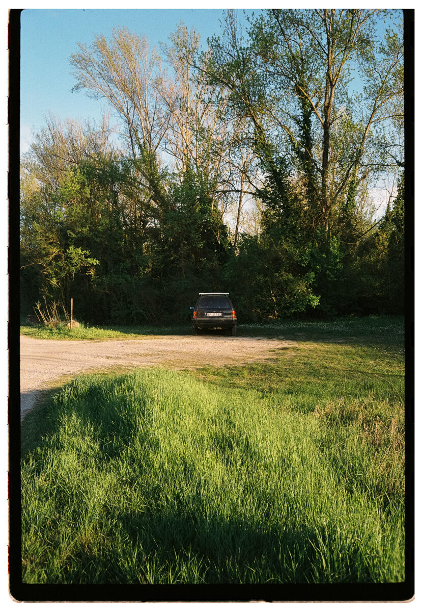 Film photograph of a dark vintage estate car parked at the end of a gravel track, surrounded by lush green spring foliage in warm late afternoon light