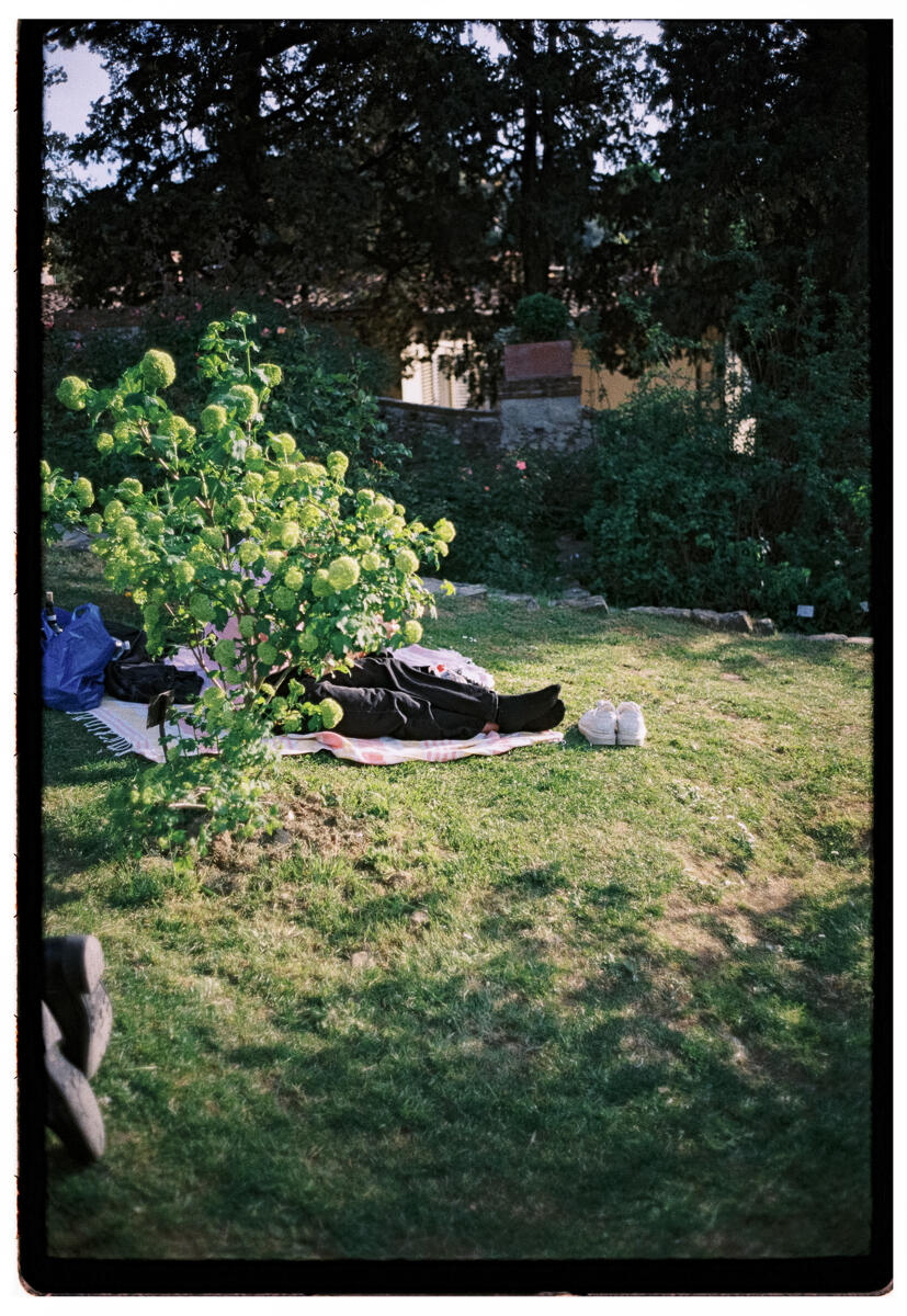 Film photograph of a person lying on a blanket in a garden, shoes placed neatly beside them, a flowering viburnum shrub in the foreground, soft afternoon light