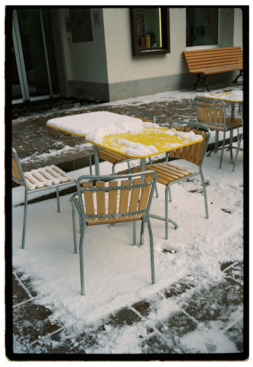 Film photograph of snow-covered outdoor café furniture, yellow table tops visible beneath fresh snow, wooden slatted chairs around them on a snowy terrace