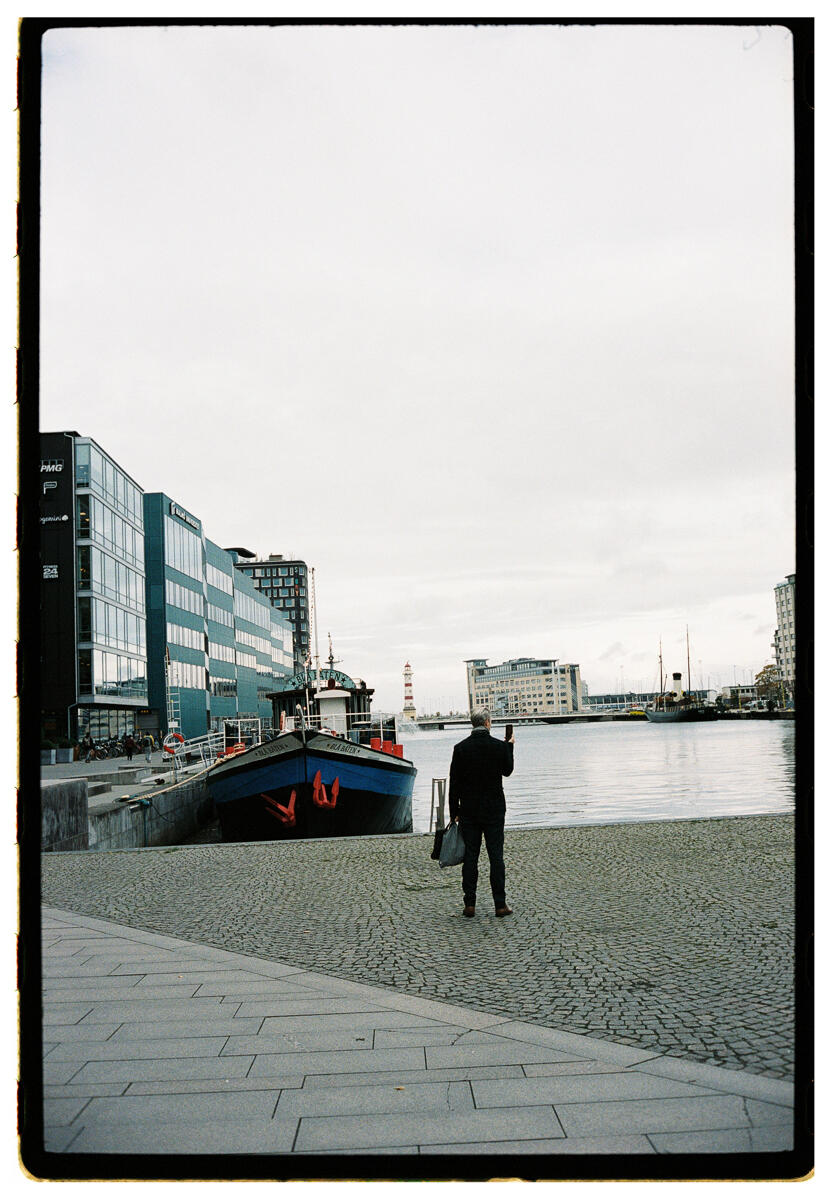 Film photograph of a lone figure walking along a harbour promenade, a blue boat moored to the left, modern glass office buildings lining the waterfront under flat grey sky
