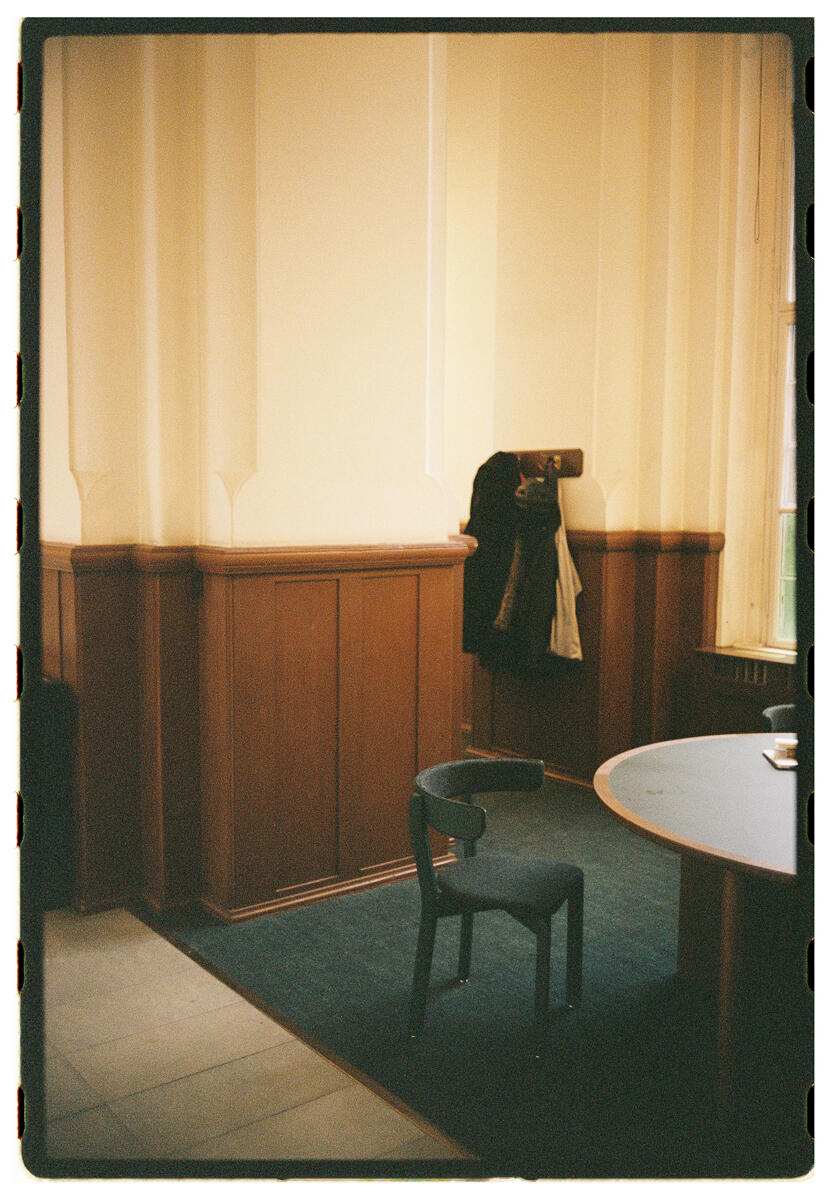 Film photograph of a quiet interior room, a single dark chair beside a round table, a coat hanging on a hook against warm yellow-cream panelled walls, natural light from a window