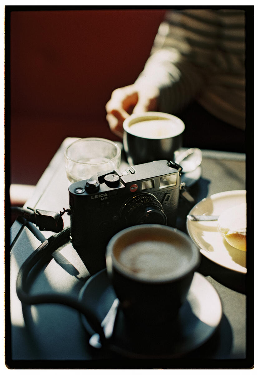Film photograph of a Leica M6 camera resting on a café table between two cups of coffee, a hand reaching for one cup in warm backlit light