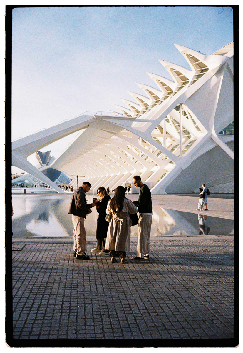 Film photograph of a group of four people gathered together in front of the Ciudad de las Artes y las Ciencias in Valencia, dramatic white architectural forms reflected in still water behind them